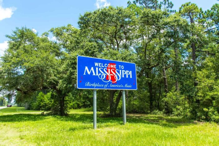 Image of a blue signboard saying Welcome to Mississippi, Birthplace of America's Music in green grass, green trees, and blue skies with clouds in the background. 