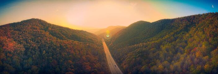 Picture of an endless road going into the sunset with lush green trees and mountains in the surroundings to represent a romantic mountain getaway. 