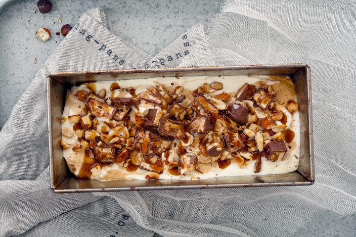 no churn salted caramel ice cream in a metallic baking loaf pan topped with pecans, snickers, and caramel