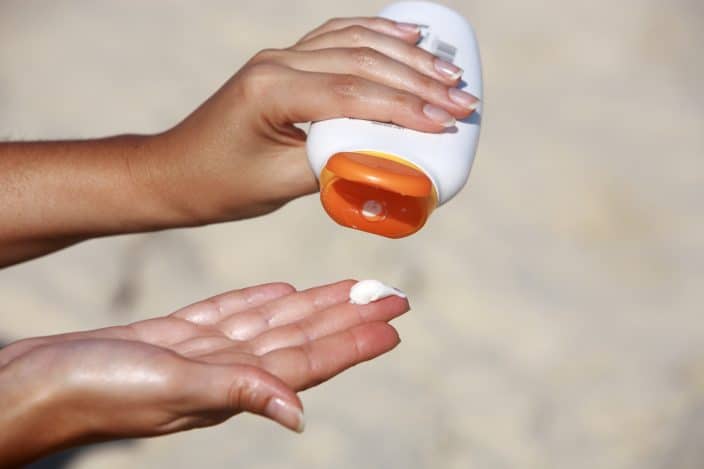 Picture of woman pouring sunscreen with an orange cap on her hands during her beach day. 