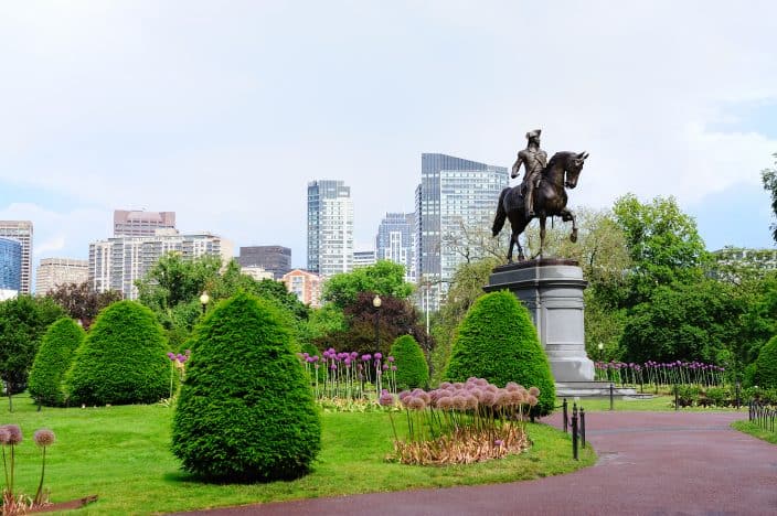 George Washington statue as the famous landmark in Boston Common Park with large purple flowers, green grass, and green shrubs with the city skyline and skyscrapers in the background in Boston, a family friendly vacation destination