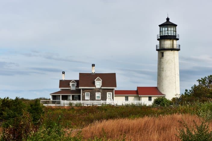 Highland Lighthouse with green and brown foliage in the front of it connected to low white house with a red and brown roof in Cape Cod, a family friendly destination