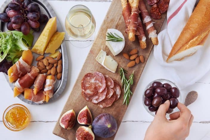 Hands getting an olive, cured meat charcuterie selection salami, chorizo, prosciutto wrapped bread sticks with fresh fig, rock melon, almonds and white wine on a white wooden table, as part of a staycation vacation
