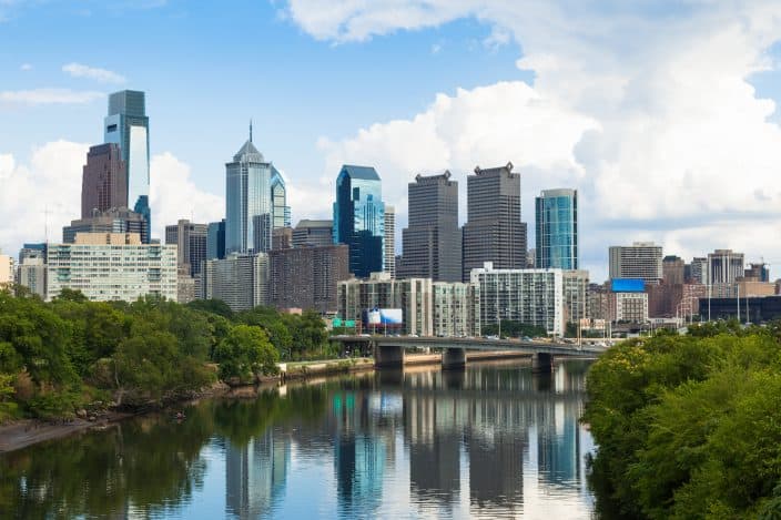 Skyline view of Philadelphia, Pennsylvania with blue skies with clods, a reflective lake with green trees and bushes around it, a great family friendly vacation destination