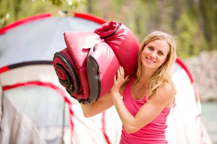 A portrait of a woman holding a red sleeping bag over her shoulder, a camping essential, with a red and silver tent in the background with green trees