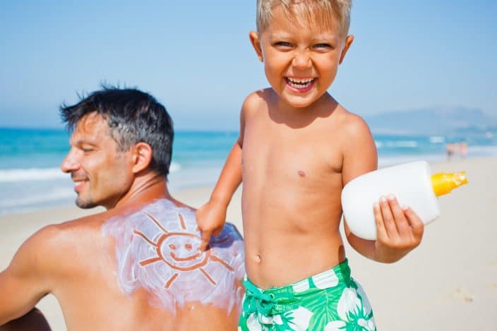 Dad and son on the beach with blue skies and waters. The father is sitting on the sand with a smiley face drawn in sunscreen on his back by the son. The son is holding a bottle of sunscreen, a camping essential. 