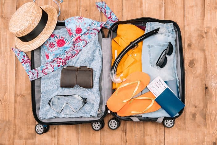 an open suitcase with traveling gear for your summer bucket list with accessories such as hat, sunglasses, flip flops, swimming gear, a t-shirt, and passport on a wooden floor