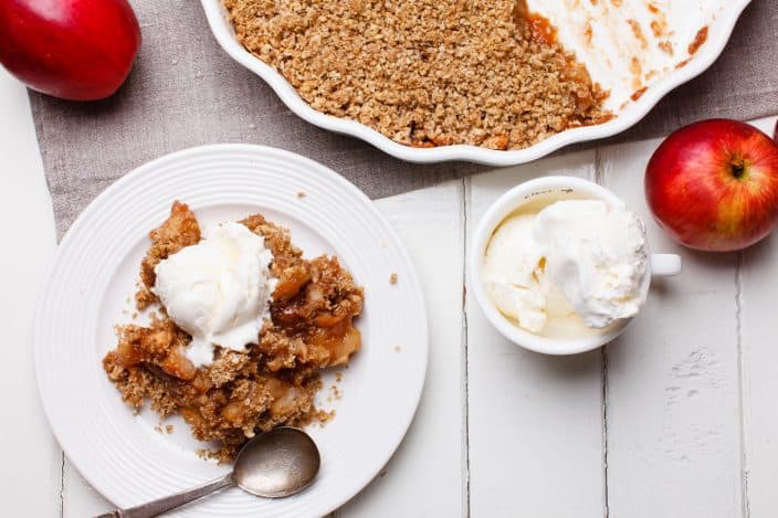 apple crisp on a plate with whipped cream on top on a white wooden background with red apples, vanilla ice cream, and a pie dish with more apple crisp