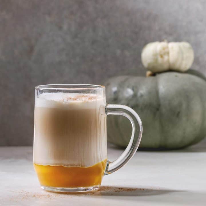 Glass of pumpkin layered spice latte with pumpkin puree, milk foam and cinnamon standing with decorative white pumpkins on white marble table with grey wall at background. Square image