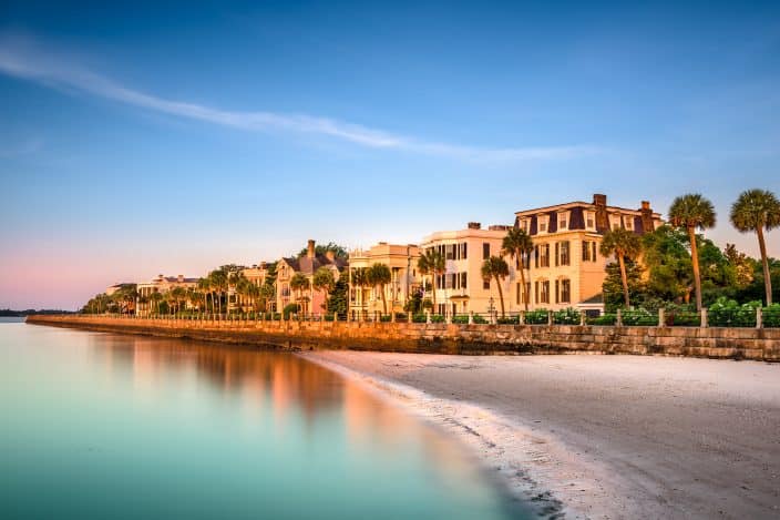 Charleston, South Carolina pastel historic houses along the coastline during sunset with blue skies and teal water and palm trees along the houses 