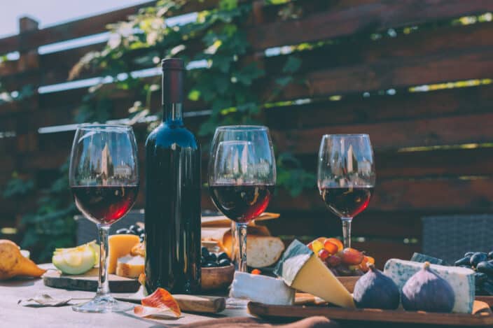 3 wine glasses filled with red wine, a wine bottle, and a cheese board with cheeses and fruits on a wooden table outside with greenery behind it. 