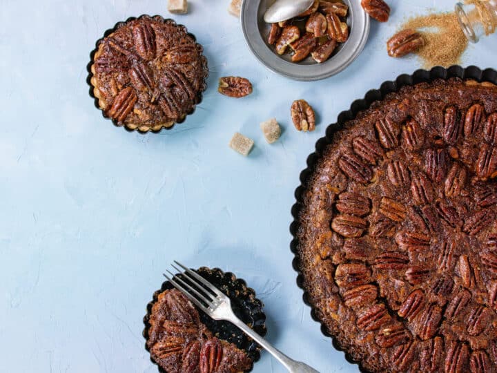 Homemade Big round maple pecan pie and small tartlets in black iron forms, served with brown sugar, maple sauce and vintage cutlery over blue textured background.
