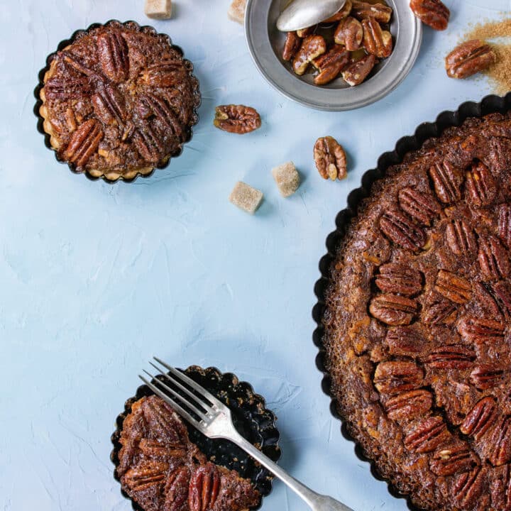 Homemade Big round maple pecan pie and small tartlets in black iron forms, served with brown sugar, maple sauce and vintage cutlery over blue textured background.