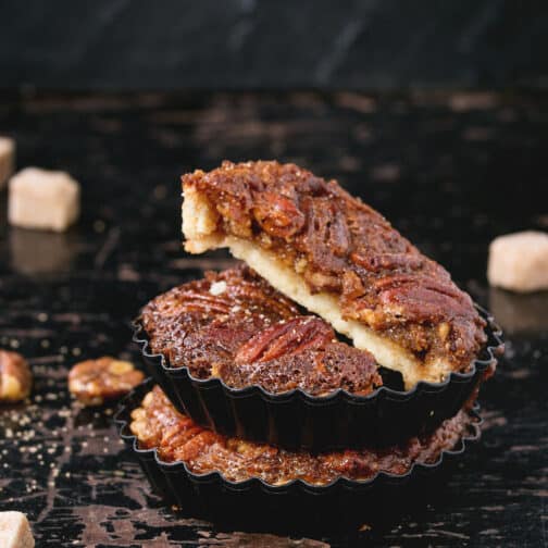 Homemade whole and half of maple pecan pie tartlets in black iron forms, served with brown sugar over old black wooden background. Square image