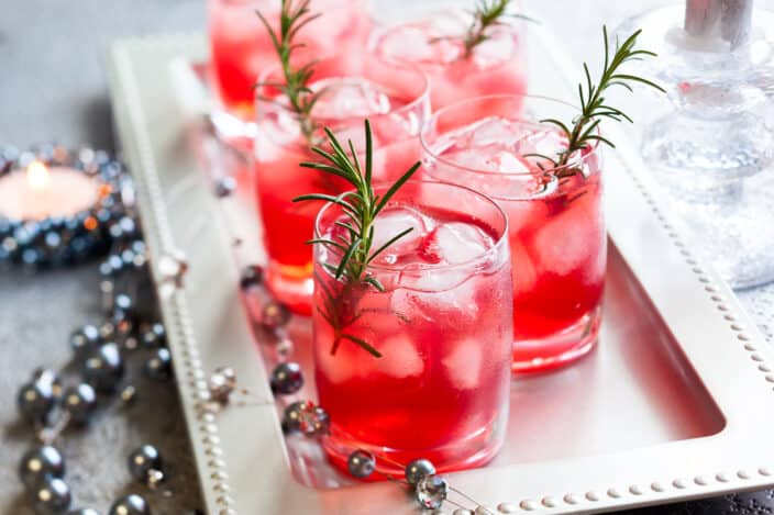 a platter with 4 glasses filled with a red cocktail with rosemary garnish and festive silver beads. An example of recipes for holiday cocktails