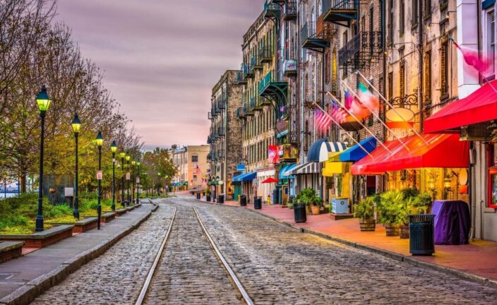a street view of Savannah, Georgia with brick buildings on the side of the road lit up by lights and flags hanging off of them and sunset lighting, an example of one of the best vacations in the south for couples