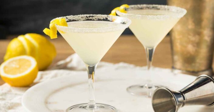 two lemon drop martinis on a table with sugar rims and a jigger and lemons lying next to them
