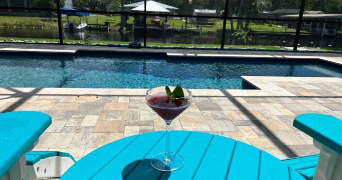 a blueberry lemon drop martini on teal table outside on the patio with a pool and canal outside with trees and blue skies