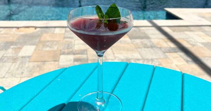 a blueberry lemon drop martini on a teal table with a pool deck and pool in the background