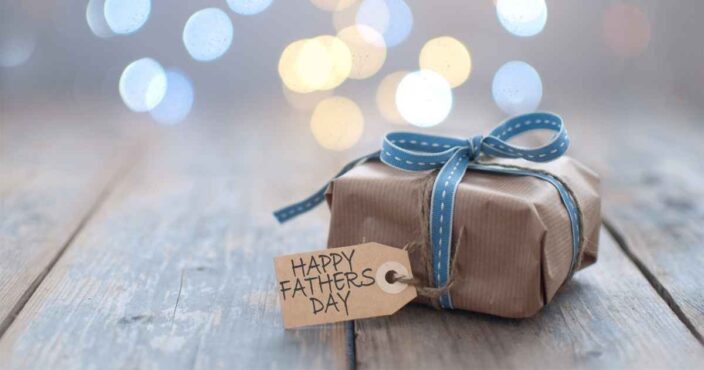 a present wrapped in brown paper on a wooden floor with twinkly lights in the background with a gift tag that says Happy Father's Day to represent a Father's Day gift guide