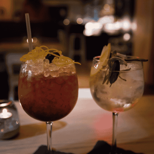 two cocktails in large wine glasses with crushed ice, one cocktail is red with orange peel on top and the other is green with green herbs and limes