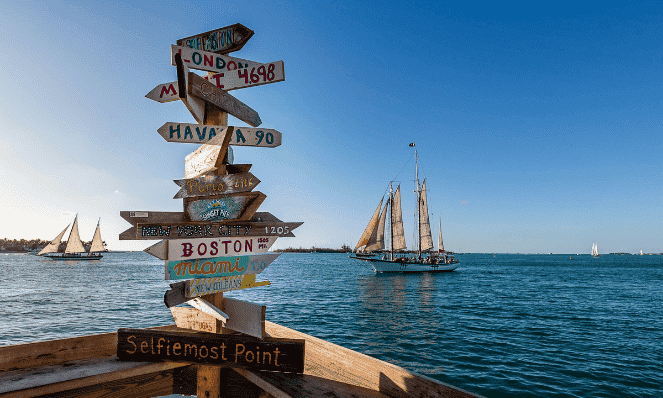 a wooden sign with multiple locations on it on a wooden dock with blue water and old sail boats in the background in Key West, an example of what you can see during a visit to Florida for vacation