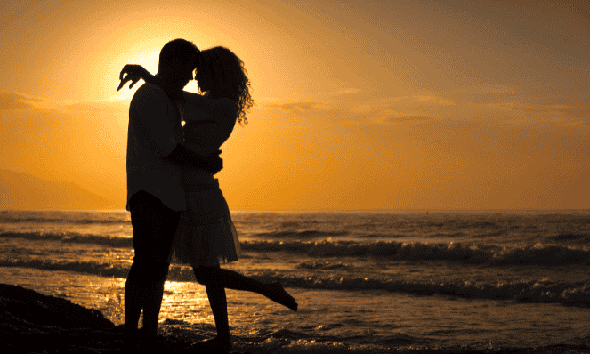 a man and a woman hugging on the beach during sunset with the ocean in the background