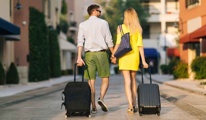 a man and a woman holding hands and walking down the street with rolling suitcases behind them to represent packing for summer vacation
