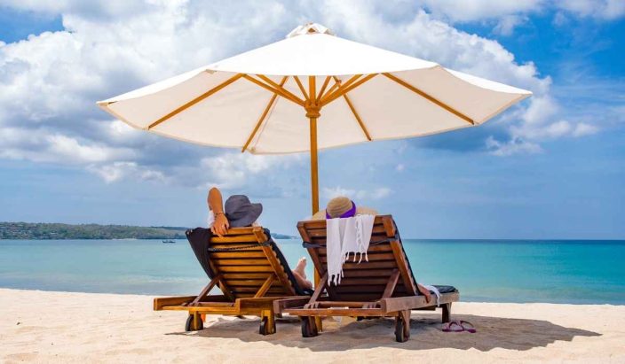 Two people with sun hats on laying on wooden lounge chairs on the beach with blue water in front of them with a large white umbrella over them and blue skies to represent summer travel