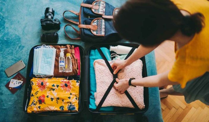 a woman packing a suitcase on a blue bed filled with a camera, floral shirt, masks, hand sanitizer, a phone and travel accessories to represent a summer packing list for international travel