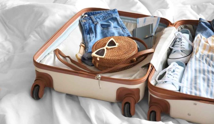 a beige and brown suitcase open on a white bed filled with a brown bag, jean shorts, a watch, ticked, blue sneakers and a striped blue shirt to represent a summer packing list for women