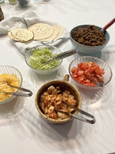 a counter with topping for tacos with ground beef including kimchi, tomatoes, shredded cheese, shredded lettuce, and flour tortillas