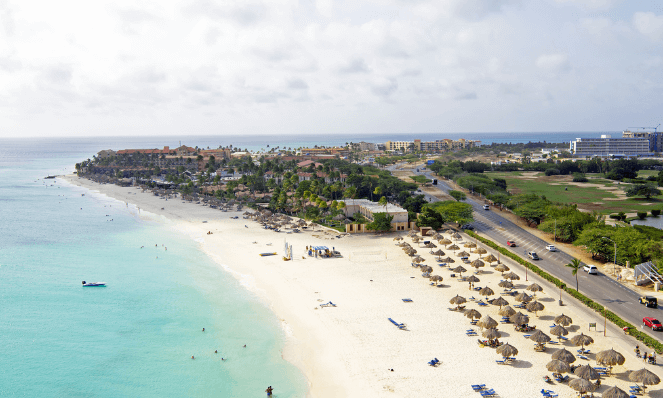 An aerial view of Manchebo beach, an aruba beach with beach huts, blue water, white sand, a town, and grey sky
